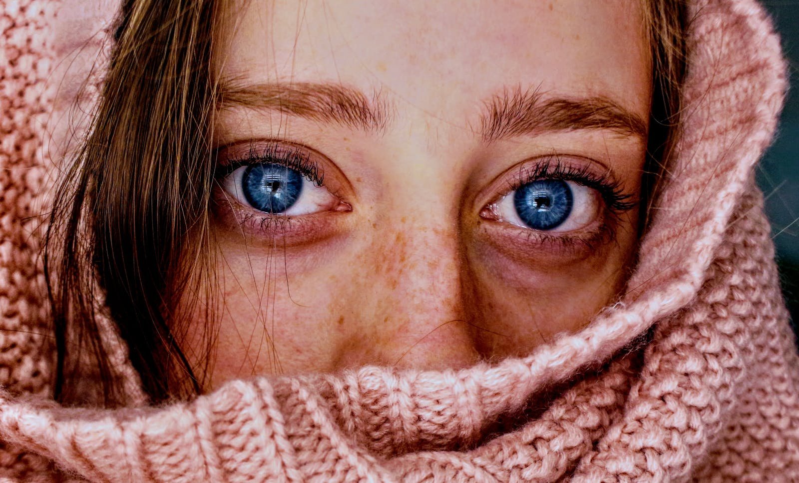 Photo by Jenna Hamra Intense close-up of a woman's blue eyes wrapped in a soft, pink knit scarf.