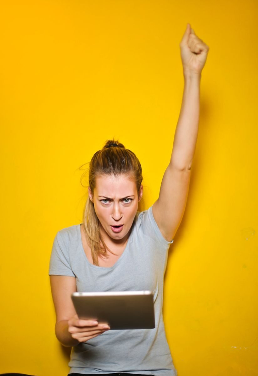 Joyful woman raises arm in celebration while holding a tablet against a vibrant yellow backdrop.